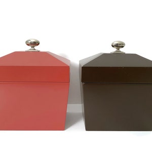 Puede incluir: Dos cajas cuadradas de madera con tapas. La caja de la izquierda es roja y la caja de la derecha es marrón. Ambas cajas tienen un pomo plateado en la parte superior.