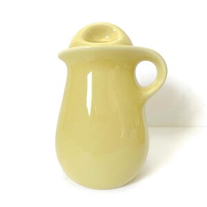 Puede incluir: Una jarra de cerámica de color amarillo pálido con asa y una pequeña tapa redondeada. La jarra tiene un acabado liso y brillante y una forma clásica y redondeada. El asa es un simple lazo y la tapa es un pequeño disco elevado.