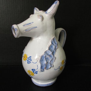 May include: A white ceramic pitcher with a pig-like face, featuring a spout, ears, and a handle. The pitcher is decorated with blue and yellow floral patterns and blue accents. The base is approximately 4 inches in diameter.