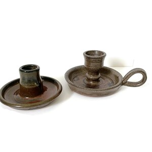 Puede incluir: Dos candelabros de cerámica. Uno es un simple candelabro redondo marrón y negro. El otro es un candelabro marrón y negro con asa y un centro elevado para la vela.