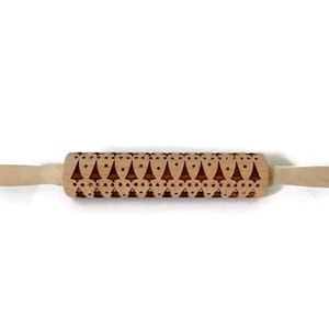 May include: A wooden rolling pin with a decorative pattern of Christmas trees and stars. The rolling pin is made of light-colored wood and features a detailed, engraved design across its rolling surface. The handles are smooth and rounded.
