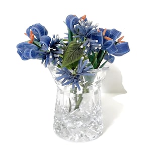 May include: A clear crystal vase filled with a bouquet of blue glass flowers with orange accents and green leaves. The vase has a cut-glass design, and the flowers are arranged to create a decorative display.