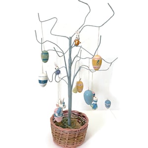 May include: A decorative Easter tree with hanging painted eggs and bunny ornaments. The tree is light blue with a woven basket base. The eggs are various colors, including blue, yellow, and green. A charming Easter decoration.