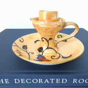 Pode incluir: Um castiçal de cerâmica vintage com um esmalte dourado e pêssego. O castiçal apresenta um design floral preto e dourado com um pássaro. O castiçal tem um pequeno prato para recolher as gotas de cera.