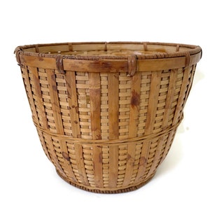 May include: A woven bamboo basket in a natural light brown hue. The basket has a flared shape and a sturdy rim and base. The design features a traditional weaving pattern. Ideal for storage or display, this basket is a practical and decorative item.