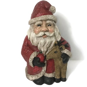 Può includere: Statuina dettagliata di Babbo Natale in abito e cappello rossi con finiture bianche. Tiene in mano una mela rossa e una piccola renna marrone. La statuina ha una finitura strutturata ed è una decorazione festiva.