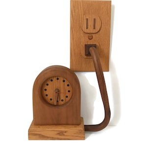 May include: A wooden clock with a round face and a cord plugged into a wooden outlet. The clock is made of light brown wood and the outlet is made of dark brown wood.