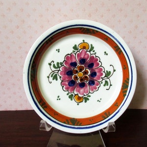 May include: A white ceramic plate with a floral design. The plate features a central flower in shades of pink, blue, and brown, surrounded by green leaves. The rim has an orange band with a blue and white border. The plate is displayed on a clear stand.