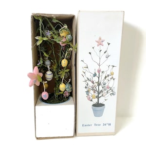 Op de afbeelding: Een Paasboom met decoratieve eieren en bloemen, gepresenteerd in een kartonnen doos. De doos leest "Easter Tree 26"H". De boom is versierd met pastelkleurige eieren en een roze ster. De boom is ongeveer 66 cm hoog.