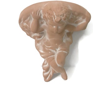 Puede incluir: Una estantería de pared con forma de querubín de color terracota. El querubín está esculpido con detalles, sosteniendo una estantería curvada. Los reflejos blancos acentúan los detalles. La estantería está diseñada para objetos decorativos.