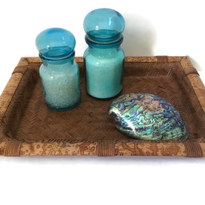May include: A rectangular woven tray with a brown border holds two blue glass jars with lids and a pearlescent abalone shell. The jars contain a white substance. The tray is on a white background.