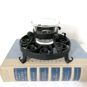 May include: A black metal candle holder with a clear glass votive cup, resting on a vintage book. The candle holder has an ornate, scrolled design and three curved feet. The book is light blue and cream-colored with the text "READER'S DIGEST CONDENSED BOOKS VOLUME 3 - 1963".