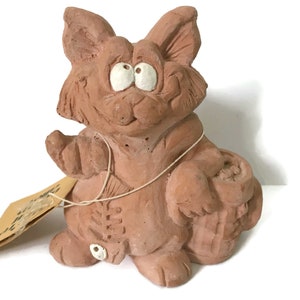 Puede incluir: Una figurita de terracota de un gato de dibujos animados con ojos grandes y un cubo. El gato sostiene un pez en su pata derecha y tiene una etiqueta atada a su pata izquierda.