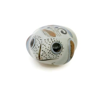 May include: A ceramic frog ornament with a white base, featuring black spots and brown, blue, and orange details. The frog has large, dark eyes and a detailed design, suitable for home decoration.