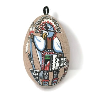May include: A hand-painted ceramic egg ornament featuring a Native American figure in traditional clothing and headdress. The figure is painted in vibrant colours, including red, blue, green, and black, against a beige background.