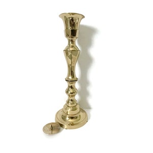 May include: A tall, gold-toned brass candlestick with a simple, elegant design. The candlestick has a round base and a single candle holder.