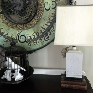May include: A white ceramic lamp with a white fabric lampshade sits on a stack of books. The lamp has a tassel on the cord. A birdcage with white porcelain birds sits on a black surface in the background.