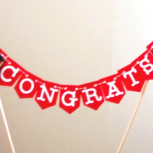 May include: A red banner cake topper with the word "CONGRATS" in white letters. The banner has small graduation caps on each end. The topper is held up by two wooden dowels with small wooden beads on top.