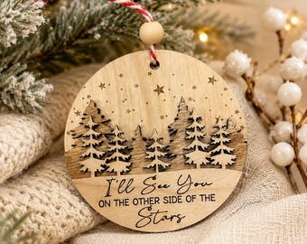 May include: A round wooden ornament featuring a laser-cut forest silhouette with layered pine trees and engraved stars. The text reads, "I'll See You ON THE OTHER SIDE OF THE Stars." It includes a red and white twine hanger with a small wooden bead.