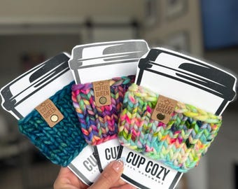 May include: Three chunky knit cup cozies in teal, multicolored, and neon pastel yarn, each attached to a white card with a coffee cup illustration and the text "COFFEE QUEEN" on a brown leather tag and "CUP COZY" printed on the card base.