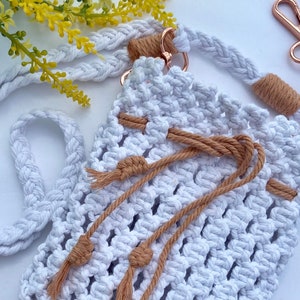Puede incluir: Bolso cruzado de macramé blanco con correa trenzada y cierre de cordón. El bolso presenta detalles en color beige y herrajes en oro rosa. Incluye un llavero a juego. Flores amarillas en el fondo.