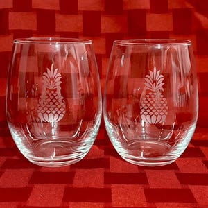 May include: Two clear stemless wine glasses with etched pineapple designs. The glasses are oval-shaped and sit on a red checkered surface. The pineapple design is centered on each glass.