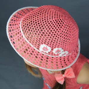 Puede incluir: Un sombrero de sol de crochet rosa con flores de crochet blancas alrededor del borde. El sombrero lo lleva una persona con el pelo largo casta&ntilde;o y un vestido rosa.