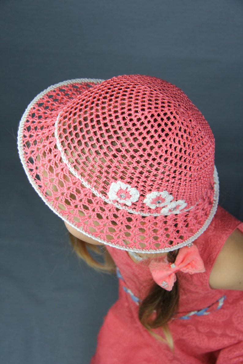 Puede incluir: Un sombrero de sol de crochet rosa con flores de crochet blancas alrededor del borde. El sombrero lo lleva una persona con el pelo largo casta&ntilde;o y un vestido rosa.