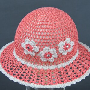 Puede incluir: Un sombrero de sol de crochet rosa con flores de crochet blancas y pedrer&iacute;a.
