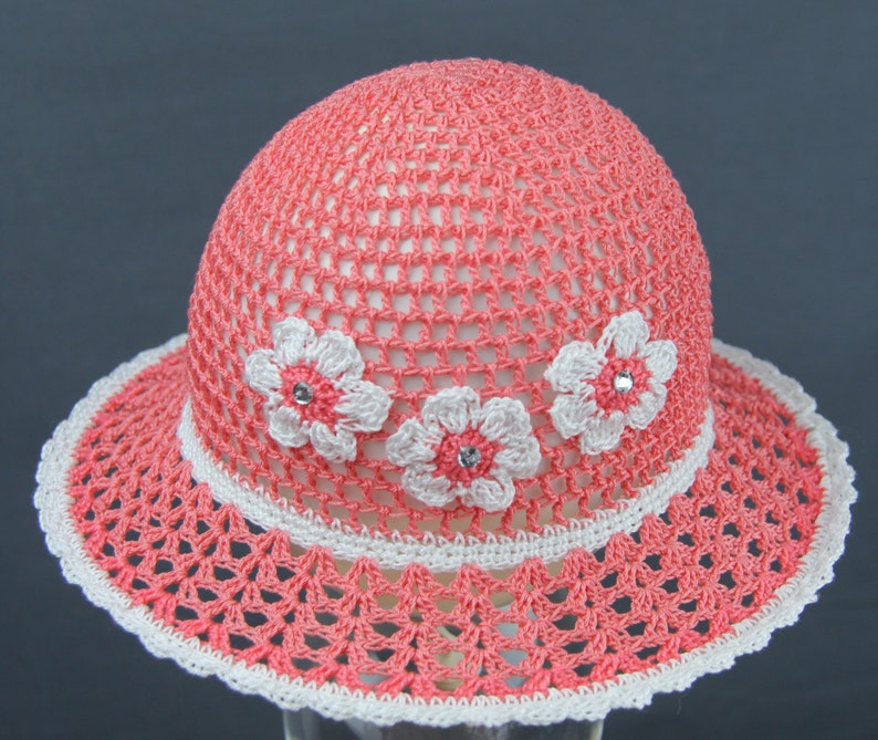 Puede incluir: Un sombrero de sol de crochet rosa con flores de crochet blancas y pedrer&iacute;a.