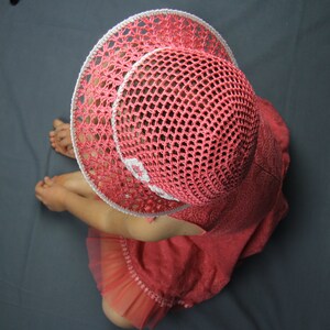 Puede incluir: Una ni&ntilde;a peque&ntilde;a lleva un sombrero de sol de crochet rosa con ribete blanco. El sombrero tiene un ala ancha y un delicado dise&ntilde;o de crochet. La ni&ntilde;a lleva un vestido rosa con un dobladillo con volantes.