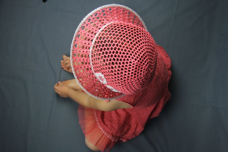 Puede incluir: Una ni&ntilde;a peque&ntilde;a lleva un sombrero de sol de crochet rosa con ribete blanco. El sombrero tiene un ala ancha y un delicado dise&ntilde;o de crochet. La ni&ntilde;a lleva un vestido rosa con un dobladillo con volantes.