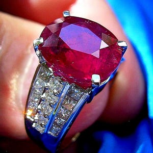 May include: A close-up of a ruby ring. The ring features a large, faceted, oval-cut ruby gemstone, set in a silver band. The band is adorned with multiple smaller, square-cut diamonds. The ring is held against a blurred background.
