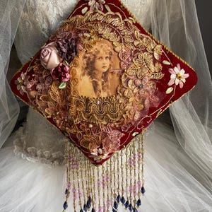 Könnte beinhalten: Ein dekoratives, rautenförmiges rotes Samtkissen mit einem Vintage-Porträt eines jungen Mädchens. Das Kissen ist mit goldener Stickerei, floralen Akzenten und Perlenfransen verziert. Eine goldene Kordel ist zum Aufhängen befestigt.