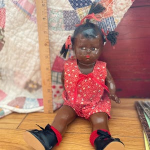 May include: A dark-skinned doll with black hair in pigtails, wearing a red romper with a white pattern and black shoes with red socks. A wooden ruler is positioned vertically next to the doll, showing a height of approximately 7.5 inches.