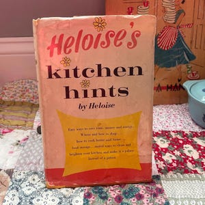 May include: A vintage cookbook, "Heloise's Kitchen Hints", with a distressed cover. The title is in a retro font with floral details. The book promises tips on saving time and money in the kitchen. It rests on a patchwork quilt, evoking a sense of nostalgia.