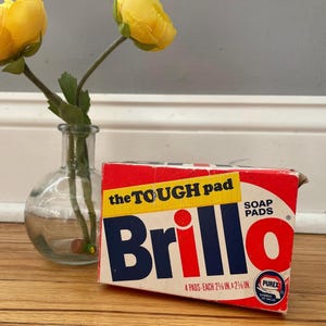 Könnte beinhalten: Eine Vintage-Schachtel mit Brillo-Scheuerschwämmen mit rotem, weißem und blauem Branding. Auf der Schachtel steht "the TOUGH pad" und "4 Pads je 6 cm x 6 cm". Im Hintergrund eine kleine Glasvase mit gelben Blumen.