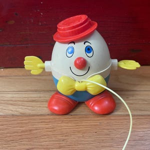 May include: A vintage toy shaped like an egg with a red hat, red feet, and yellow hands and bow tie. The egg has a smiling face with blue eyes and a red nose. A yellow string extends from the toy.