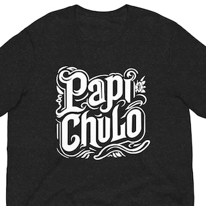 May include: Dark gray t-shirt with the words "Papi Chulo" in a white, ornate font. The text is centered on the shirt and features decorative flourishes and a small floral element. The shirt is made of a soft material.