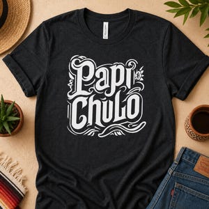 May include: Dark gray t-shirt with the words "Papi Chulo" in white, stylized lettering. The shirt is folded, with rolled-up sleeves. A straw hat, succulent plant, and jeans are visible in the background.