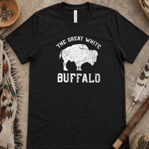 Puede incluir: Camiseta negra con un gráfico blanco de un búfalo y el texto "THE GREAT WHITE BUFFALO" en una fuente desgastada. La camiseta es de cuello redondo y tiene mangas cortas.