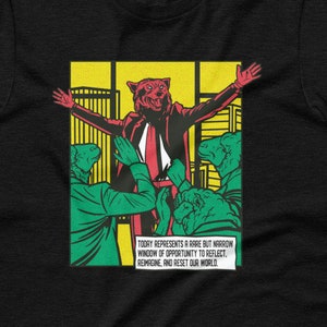 May include: Black t-shirt with a comic book style graphic of a red wolf in a suit with the text "TOORY REPRESENTS A RARE BUT NARROW WINDOW OF OPPORTUNITY TO REFLECT, REMAGNE, AND RESET OUR WORLD."