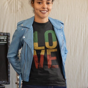 May include: A woman wearing a black t-shirt with the word "LOVE" in colourful letters, a blue denim jacket, and blue jeans. She is standing in a music studio with a guitar, amplifier, and other musical equipment in the background.