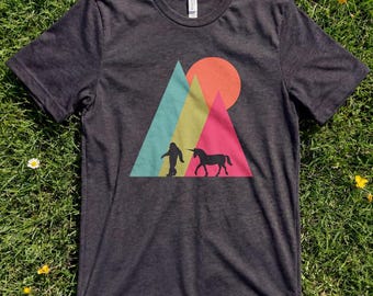 May include: Dark gray t-shirt featuring a colorful graphic of a mountain scene with a Bigfoot silhouette and a unicorn. The design includes a sun and triangular mountains in teal, yellow, and pink. A casual tee for outdoor enthusiasts.