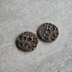 以下が含まれることがあります： 複雑な透かし彫りのデザインが施された、2つのアンティークブロンズ色のボタン。円形のボタンは、織り交ぜられた線と形状の精巧なパターンを持ち、テクスチャード加工された外観を作り出しています。灰色の表面に配置されています。