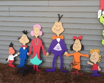 May include: A collection of wooden cutouts depicting a family, each with distinct colours and designs. The figures are holding hands, with the tallest wearing a purple suit and orange bow tie. They stand on a bed of brown mulch.