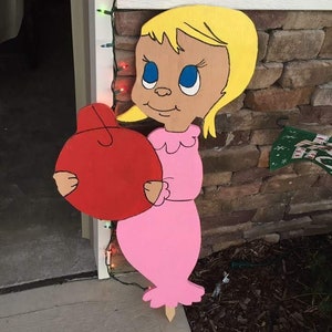Può includere: Decorazione da esterno in legno raffigurante un personaggio dei cartoni animati con capelli gialli, occhi blu e un vestito rosa, che tiene in mano un ornamento rosso. La decorazione è dipinta con colori vivaci e ha un paletto per il terreno.
