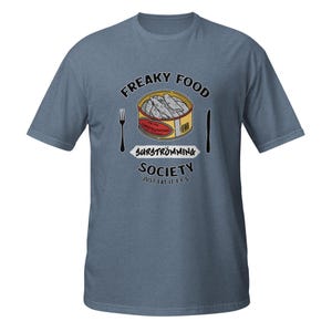 May include: Blue t-shirt with a graphic featuring a can of surströmming, a Swedish fermented herring dish. The design includes the words "Freaky Food Society" and "Just Eat It F.F.S.", along with a fork and knife. The can is yellow with a red label.