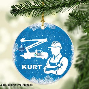 May include: Round blue Christmas ornament with a white silhouette of a utility truck, a worker, and the name "KURT." The ornament has a snowflake design and the Duke Energy logo. A gold string is attached for hanging.