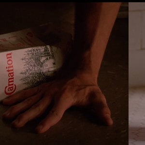 May include: A white carton of Carnation milk is lying on the floor, spilled and partially open. The carton is next to a white milk carton with a missing child poster on it. The poster features a photo of a young boy with brown hair and a black hooded sweatshirt. The poster reads "MISSING" and "LADDIE THOMPSON".